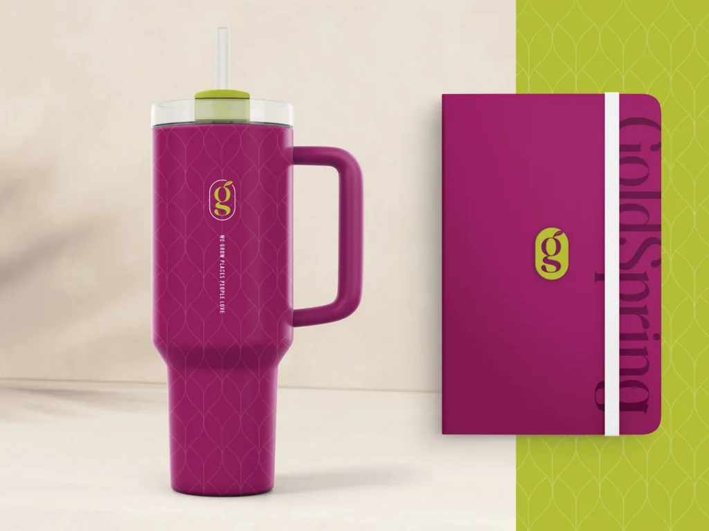 GoldSpring Marketing branded magenta tumbler with the "We grow places people love" tagline and leaf pattern, next to a magenta notebook with a lime green monogram.